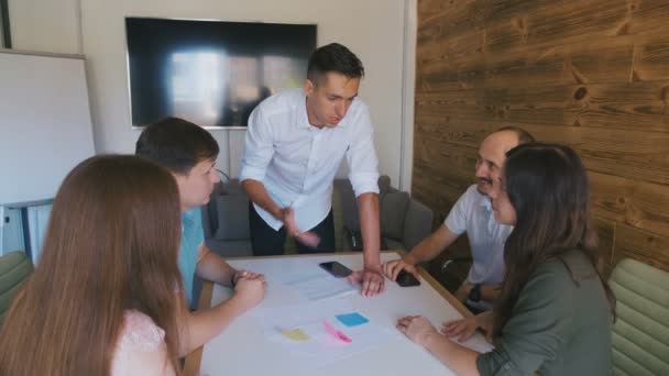 Бізнес команда обговорює проект за великим офісним столом. Чоловічий лідер команди стоїть біля столу і дає керівництво молодій творчій команді . — стокове відео