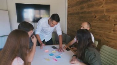 İş Ekibi Büyük Ofis Tablosunda Bir Projeyi Tartışıyor. Erkek Takım Lideri Masa'nın yanında duruyor ve genç yaratıcı ekibe Yön veriyor.