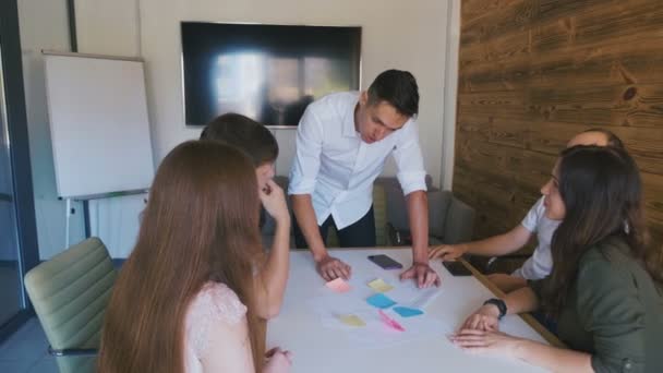 Business team bespreekt een project aan een grote kantoor tafel. Mannelijke teamleider staat in de buurt van tafel en geeft leiding aan jong creatief team. — Stockvideo