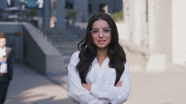 Mladá krásná obchodní žena v brýlích, stojící nedaleko moderní kancelářské budovy, dívá se na kameru a usmívá se. Obchodní lidé, kteří chodí po pozadí. — Stock video