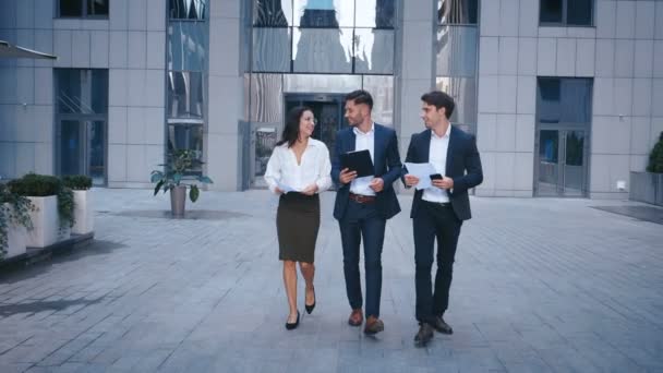 Des gens d'affaires prospères Discutez d'affaires et marchez dans le quartier central des affaires. Deux jeunes hommes et une femme en costume Communiquer et marcher près de l'immeuble de bureaux du centre d'affaires . — Video