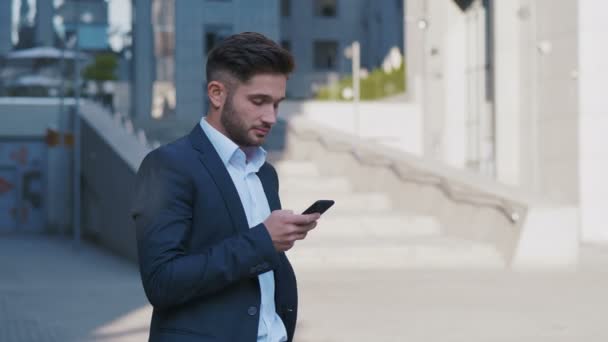 Mladý pohledný obchodník stojící poblíž budovy Big Office. Zadání zprávy na jeho zařízení smartphone. vousatý muž na klasické skafandru. Obchodní životní styl. — Stock video