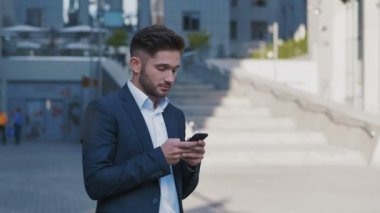 Genç Yakışıklı İşadamı Büyük Ofis Binası'nın yanında duruyor. Onun Smartphone.Bearded Man Klasik Takım Giyen Bir Mesaj Yazma. İş Yaşam Tarzı.
