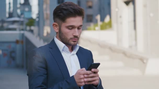 Νέος όμορφος επιχειρηματίας στέκεται κοντά στο μεγάλο κτίριο γραφείων. Δακτυλογράφηση ενός μηνύματος στο smartphone του. Επιχειρηματικός τρόπος ζωής. — Αρχείο Βίντεο