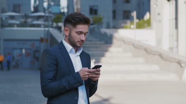 Junger gutaussehender Geschäftsmann, der in der Nähe eines großen Bürogebäudes steht. Er tippt eine Nachricht auf sein Smartphone, ein bärtiger Mann im klassischen Anzug. Geschäftsleben. — Stockvideo