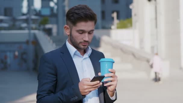 Hombre joven SMS mensajes de texto utilizando la aplicación en el teléfono inteligente en la ciudad cerca de edificio de oficinas. Guapo joven hombre de negocios beber café con teléfono inteligente — Vídeos de Stock