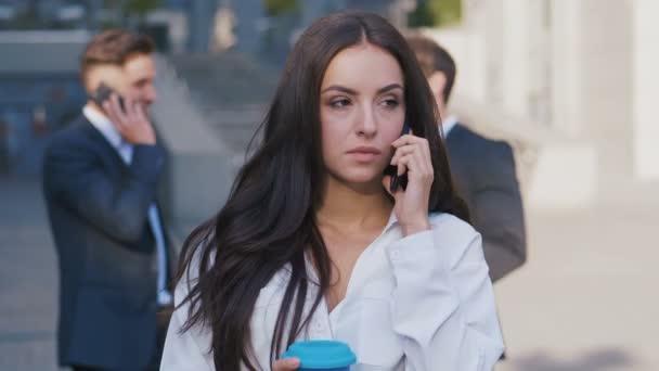 Schöne Geschäftsfrau telefoniert während der Kaffeepause auf den Straßen des Geschäftsviertels in der Nähe des Bürogebäudes Business Center und einer Gruppe von Geschäftsleuten im Hintergrund. — Stockvideo