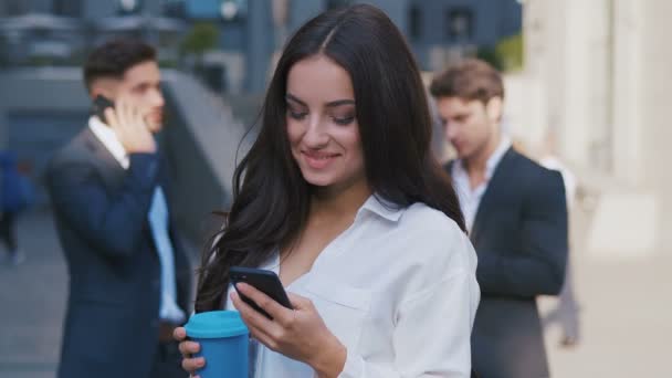 Портрет усміхнена молода жінка Бізнесжінка за допомогою смартфона біля офісної будівлі на каву розбийте на вулиці ділового району. Група зайнятих ділових людей на задньому плані. — стокове відео