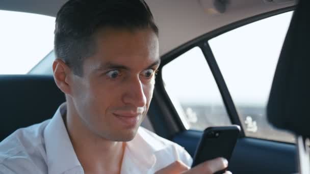 ビジネスマンは、スマートフォン上で良いメッセージを読んで、彼は幸せで笑顔です。男は運転手やタクシーで車に乗り、携帯電話を使用します — ストック動画