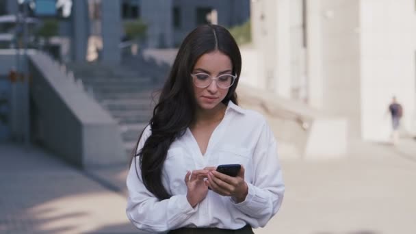年轻的美丽的商务女性戴着眼镜和白色衬衫使用智能手机，站在办公楼商务中心附近. — 图库视频影像