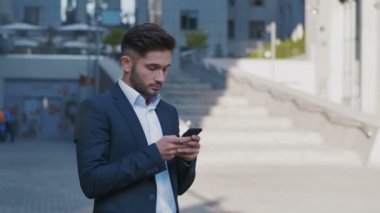 Genç Yakışıklı İşadamı Büyük Ofis Binası'nın yanında duruyor. Onun Smartphone.Bearded Man Klasik Takım Giyen Bir Mesaj Yazma. İş Yaşam Tarzı.
