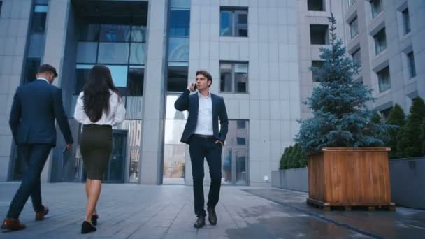 Udane biznesmen chodzenie na ulicach dzielnicy biznesowej i rozmawiać na telefon komórkowy. Przekonany, atrakcyjny kaukaski mężczyzna w garnitur chodzenie na zewnątrz w pobliżu dużego biurowego budynku i za pomocą smartfona. — Wideo stockowe