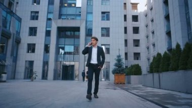 Başarılı İş Adamı İş Mahallesi Sokaklarında Yürüyor ve Cep Telefonuyla Konuşuyor. Büyük ofis binasının yakınında yürüyen ve akıllı telefon kullanan takım elbiseli kendine güvenen çekici beyaz adam.