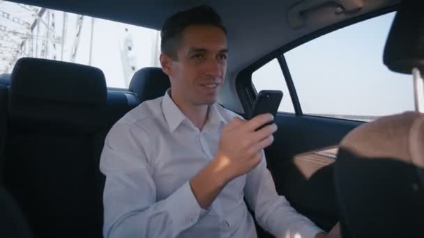 年轻的商人使用他的智能手机在旅行与司机的汽车。男子正在检查邮件，聊天或新的在线，而他乘坐出租车 — 图库视频影像