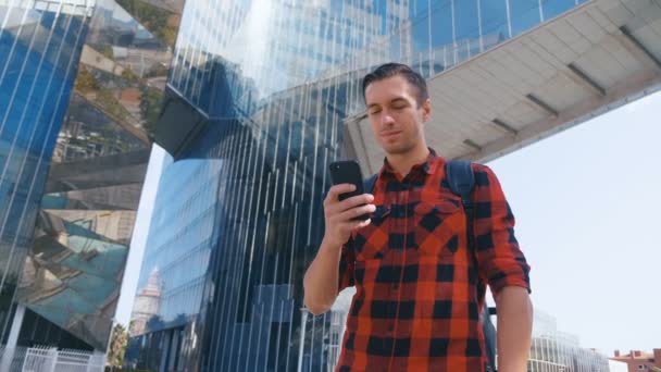 Όμορφος Καυκάσιος άνθρωπος με το καρό πουκάμισο και με ένα σακίδιο συνομιλίας και περιήγηση στο κινητό τηλέφωνο κοντά στο μοντέρνο γυαλί κτίριο. — Αρχείο Βίντεο
