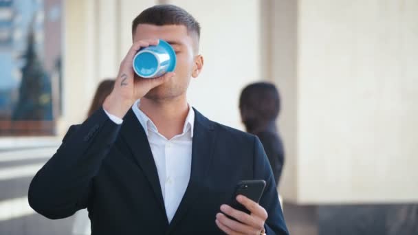 Добре одягнений підприємець стоїть біля свого офісу. Пити каву, щоб піти. Набирає повідомлення на свій смартфон. Люди спілкуються на фоні. Діловий район . — стокове відео