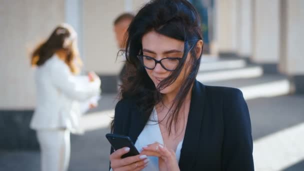 Primer plano de la hermosa joven mujer de negocios con mensajero de mensajes de texto de teléfonos inteligentes y sonriendo fuera mientras trabaja en el centro de negocios . — Vídeos de Stock