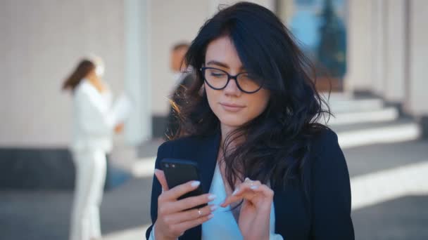 Молодая предпринимательница в очках, стоящая в городе со смартфоном в руках. Выглядит счастливым и довольным. Офисное здание на заднем плане . — стоковое видео