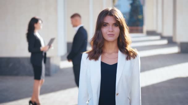 Portret van Pretty Young Businesswoman Staande in de stad in de buurt van Office Building. Je ziet er gelukkig en tevreden uit. Witte jas gekleed. Zakelijke mensen die op de achtergrond communiceren. — Stockvideo