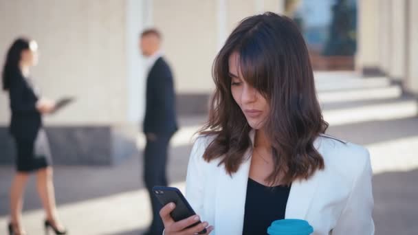 Porträt einer attraktiven Geschäftsfrau, die in der Kaffeepause ihr Smartphone benutzt. schöne Managerin steht draußen und schreibt SMS auf ihr Smartphone in der Nähe von Bürogebäuden Business Center. — Stockvideo