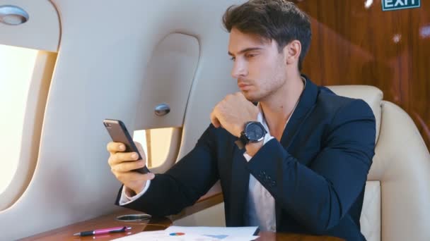 Młody przystojny dyrektor bankowy biznesmen używający smartfona podczas lotu samolotem. Udany biznesmen podróżujący swoim prywatnym odrzutowcem. — Wideo stockowe