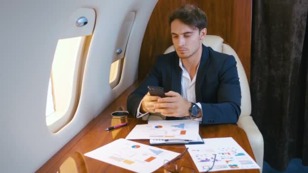 飛行機の中で飛行中にスマートフォンを使用して働く若いハンサムなチーフ銀行家取締役ビジネスマン。民間のジェット飛行機に乗って成功したビジネスマン. — ストック動画