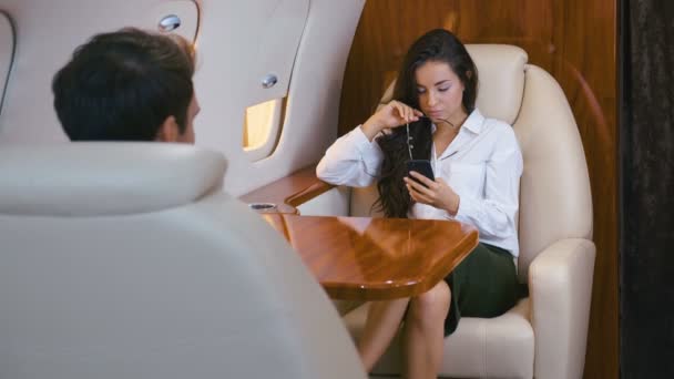 Attraktiv affärskvinna i glasögon med smartphone i händerna Resor på ett privat flygplan. Affärsresa. Klassiskt klädd. Flicka miljonär flyger i jetplan i en businessklass. — Stockvideo