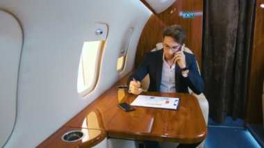 Meşgul işadamı, kişisel jet uçağıyla uçarken belgelerle çalışır ve cep telefonuyla konuşur. Uçak Business Class.