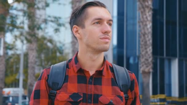 Retrato de un joven guapo con la camisa a cuadros y con una mochila mirando a la cámara. Fondo del edificio urbano de cristal grande . — Vídeos de Stock