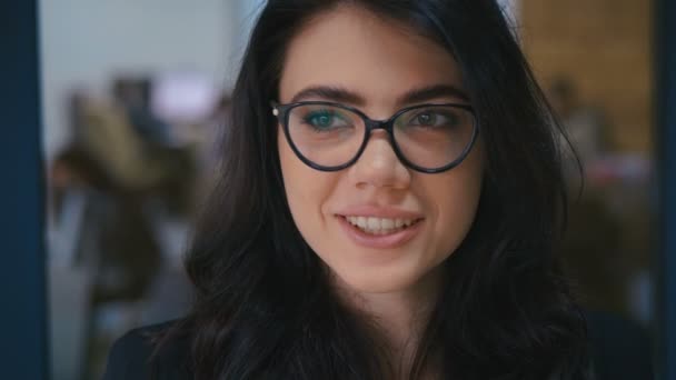 Krásná usmívající se sebevědomá mladá žena hezká tvář dívající se do kamery v kanceláři, zblízka portrét. Nosí brýle. Kancelářské práce na pozadí. — Stock video