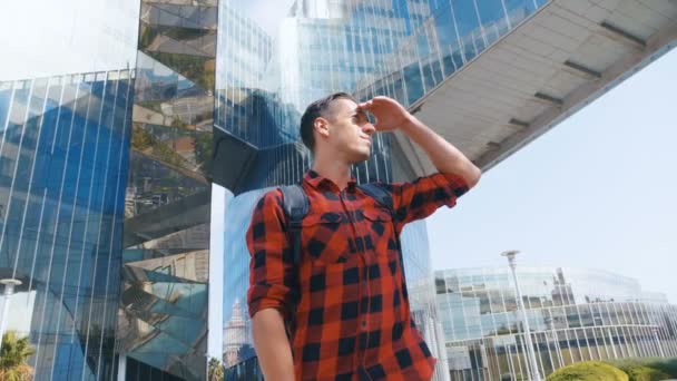 Retrato de turista jovem bonito na camisa xadrez e com uma mochila olhando ao redor e depois para a câmera. Grande vidro edifício urbano fundo . — Vídeo de Stock