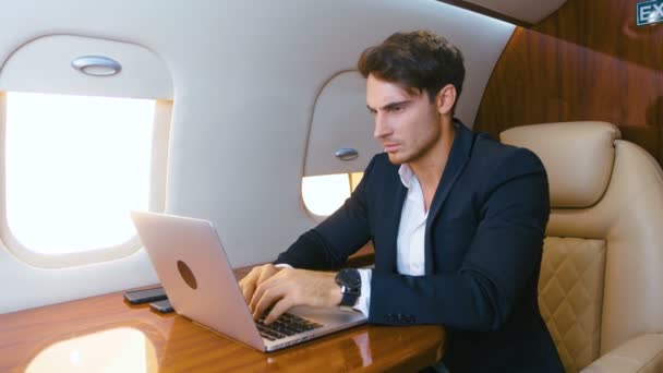 若いビジネスマンは彼のプライベート飛行機のラップトップで働いています。一流の飛行機で旅行する大成功したビジネスマン。古典的なスーツを着て実業家. — ストック動画
