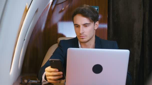 Jonge zakenman werkt op laptop en gebruikt smartphone in zijn privévliegtuig. Succesvolle zakenman op reis in eersteklas vliegtuig. Klassiek pak gekleed. Zakenmensen. — Stockvideo