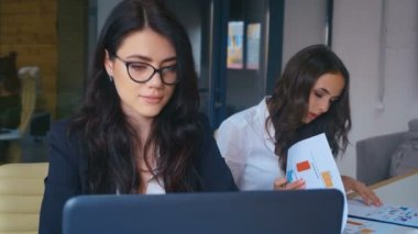İş kadını kendini mutlu hisseder gülümser ve ofiste kameraya bakar. Modern ofiste dizüstü bilgisayar kullanan genç bir kadın..