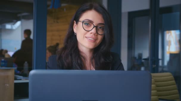 Obchodnice se cítí šťastná, usmívá se a dívá se na kameru v kanceláři. Mladá žena pracující pomocí notebooku na stole v moderní kanceláři. — Stock video