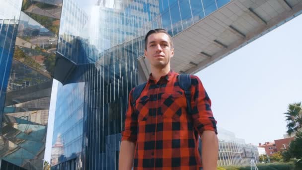 Portret przystojnego młodego mężczyzny turysty w koszuli w kratę i z plecakiem rozglądającym się dookoła, a potem przed kamerą. Duże szklane tło budynku miejskiego. — Wideo stockowe