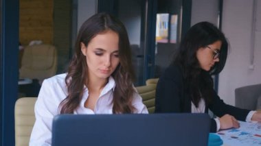 Genç ve güzel bir iş kadını, modern ofiste masa başında çalışırken gülümseyip kameraya bakarken mutlu oluyor..