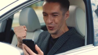 Arabada otururken hareketli görünmesine şaşırdım. Akıllı telefondan iyi haberler okuyan heyecanlı sürücünün portresi. Başarılı iş adamı dışarıda cep telefonuyla zaferi kutluyor..