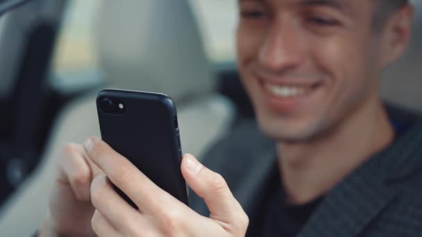 Close-up portret van lachende mannelijke zakenman zittend achter het stuur van een auto en met behulp van smartphone. — Stockvideo