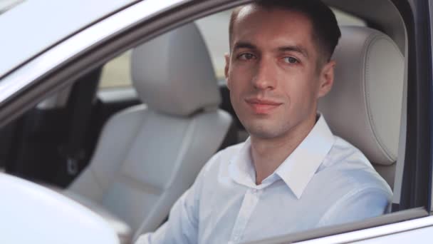 Retrato de un hombre de negocios con una camisa blanca sentado en un coche en un asiento de conductores y sonriendo . — Vídeos de Stock