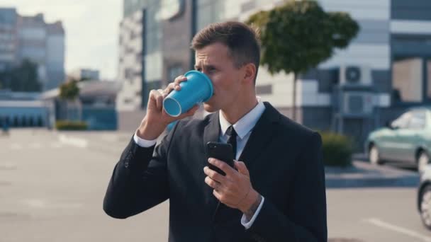 Porträt eines attraktiven Jungunternehmers, der sein Smartphone benutzt und Kaffee trinkt. In der Nähe des modernen Bürogebäudes. Im klassischen Anzug. Soziales Netzwerk — Stockvideo