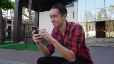 Modern ofisin yanındaki bankta otururken akıllı telefonunu kullanan yakışıklı bir gencin portresi. Ekose tişörtlü bir adam boş zaman geçirirken telefon ekranını kaydırıyor.