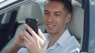 Bir arabanın direksiyonunda oturan ve akıllı telefon kullanan gülümseyen erkek işadamının yakın plan portresi..