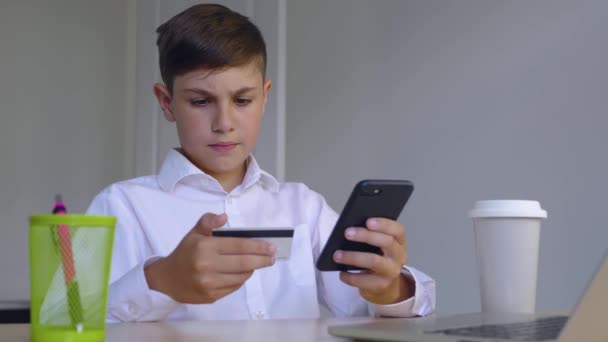 Gyönyörű fiú 10 éves otthon ülve online fizet az interneten keresztül bankkártyáról okostelefonon. A gyerek az online áruházban vásárol. Online banki szolgáltatások okostelefonnal. — Stock videók