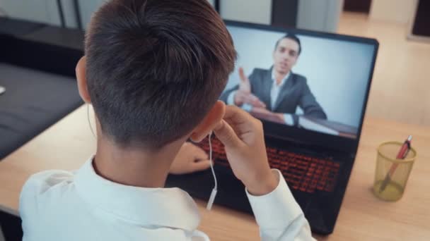 Niño en auriculares escuchando lección de la escuela en línea con el profesor joven en la pantalla del ordenador portátil. Niño sentado en el escritorio en la habitación de los niños en casa y estudiando en línea. Autoaislamiento y educación en línea . — Vídeo de stock