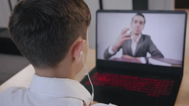 Niño en auriculares escuchando lección de la escuela en línea con el profesor joven en la pantalla del ordenador portátil. Niño sentado en el escritorio en la habitación de los niños en casa y estudiando en línea. Autoaislamiento y educación en línea . — Vídeo de stock