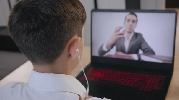 Pojke i hörlurar lyssnar online skollektion med ung manlig lärare på bärbar skärm. Grabben sitter vid skrivbordet i barnrummet hemma och studerar online. Egenisolering och online-utbildning. — Stockvideo