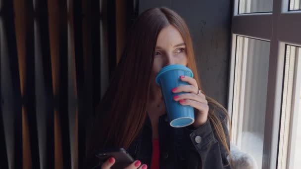 Νεαρή σκεπτική γυναίκα κάθεται στο σπίτι κοντά σε ένα παράθυρο, χρησιμοποιεί ένα smartphone και πίνει καφέ. — Αρχείο Βίντεο
