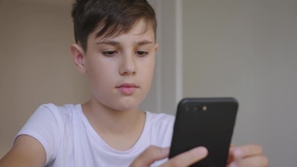 Retrato de niño guapo de 12 años usando smartphone en casa. Colegial caucásico utiliza un teléfono inteligente mientras está sentado en una mesa en casa — Vídeos de Stock