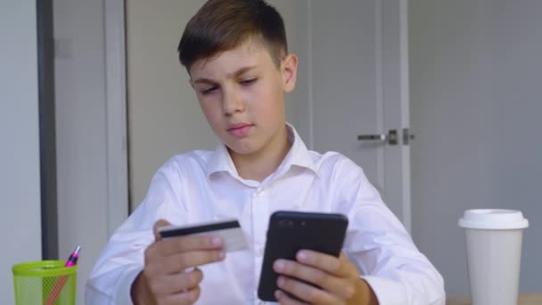 Beau garçon de 10 ans assis à la maison effectue des paiements en ligne via Internet à partir d'une carte bancaire sur smartphone. Enfant fait des achats dans la boutique en ligne. Services bancaires en ligne avec téléphone intelligent. — Video